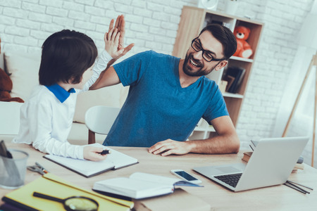 Man Spends Time with His Son. Father of Boy is Engaged in Raising Child. Father is Doing a Homework with Son. Father and Son Giving Five to Each Other. People is Sitting at Table. People at Home.の写真素材