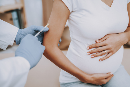 Pregnant Girl at the Gynecologist Doctor. Doctor is a Young Man. Doctor Injects a Medicine to a Woman. Girl is Sitting on Couch and Touching Her Belly. People Located at Clinic. Closeup.の写真素材