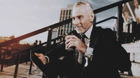 Businessman is Using a Tablet PC. Businessman is Old Smiles Man. Businessman is Holding a Coffee Cup. Man is Looking to Distance. Man in Black Suit. Businessman is Sitting on Staircase. Sunny Daytime.の写真素材