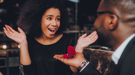 African Handsome Man Proposing to Girlfriend. Romantic Couple in Love Dating. Cutel Man and Girl in a Restaurant. Romantic Concept. Surprised Attractive Woman Getting a Marriage Proposal.の写真素材