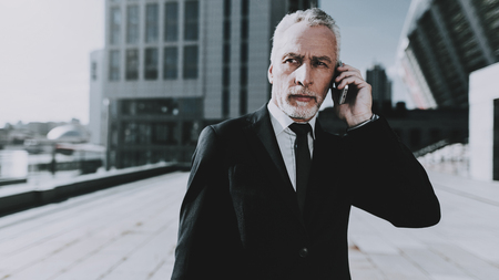 Businessman is Talking on Mobile Phone. Businessman is Old Serious Man. Man Wearing in Black Suit. Man Looking to Distance. Businessman is Standing on Roof of Skyscraper. Sunny Daytime.の写真素材