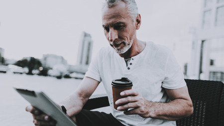 Man is Using a Tablet PC. Man is Looking on Tablet Screen. Old Smiling Man. Person is Sitting on Couch. Person is Holding a Coffee Cup. Sunny Daytime. Person Located in Business District.の写真素材