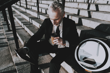 Businessman is Using a Tablet PC. Businessman is Old Smiling Man. Man is Holding a Coffee Cup. Man Wearing in Black Suit. Businessman is Sitting on Staircase. Monowheel near a Man. Sunny Daytime.の写真素材