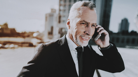 Businessman is Talking on Mobile Phone. Businessman is Old Smiling Man. Man Wearing in Black Suit. Man Looking to Distance. Businessman is Standing on Roof of Skyscraper. Sunny Daytime.の写真素材