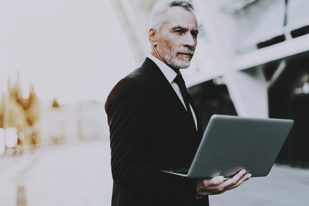 Businessman is Holding a Laptop Computer. Businessman is Old Serious Man. Man Wearing in Black Suit. Man Looking to Distance. Businessman is Standing on Roof of Skyscraper. Sunny Daytime.の写真素材