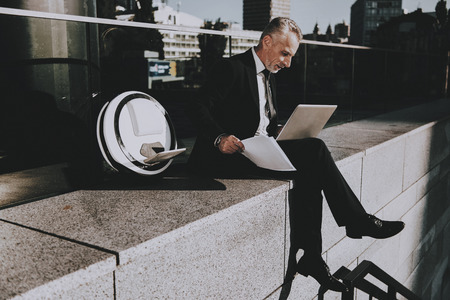 Businessman is Using a Laptop Computer. Businessman is Old Smiling Man. Man Wearing in Black Suit. Man Holding a Papers. Businessman is Sitting on Roof. Monowheel near a Man. Sunny Daytime.の写真素材