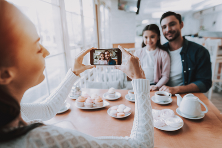 Family Resting in Cafe. Family is a Mother, Father and Daughter. Mother Photographs Father with Daughter on Mobile Phone. Persons is Sitting at Table. People is Happy and Smiling. Daytime.の写真素材