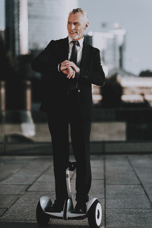 Businessman is Riding on scooter. Businessman is Looking a Time on Clock. Businessman is Old Smiling Man. Man Wearing in Black Suit. Man Looking to Distance. Businessman is Riding on Roof. Daytime.の写真素材