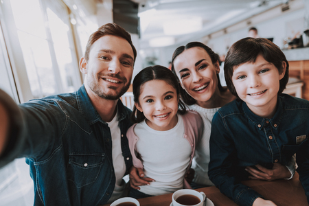 Family Resting in Cafe. Family is a Mother, Father, Daughter and Son. People is Doing Selfie on Mobile Phone. People is Happy and Smiling. Persons is Sitting at Table. Family Portrait. Sunny Daytime.の写真素材