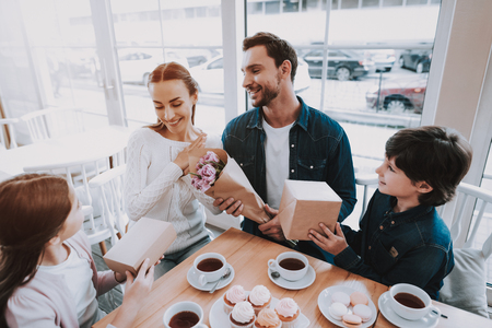 Family Celebrating a Holiday in Cafe. Family is a Mother, Father, Daughter and Son. Father and Children is Giving a Flowers and Gifts to Mother. People is Happy and Smiling. Cakes and Tea on Table.の写真素材