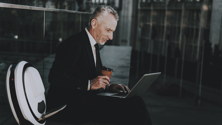 Businessman is Using a Laptop Computer. Businessman is Old Smiling Man. Man Wearing in Black Suit. Man Holding a Coffee Cup. Businessman is Sitting on Roof. Monowheel near a Man. Sunny Daytime.の写真素材