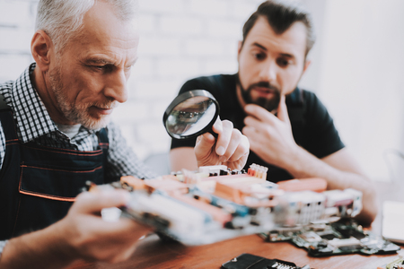 Two Men Repairing Hardware Equipment from PC. Repair Shop. Worker with Tools. Computer Hardware. Young and Old Workers. Modern Devices. Digital Device. Laptop on Desk. Electronic Devices Concept.の写真素材