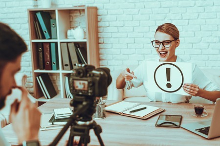 Blogger Makes a Video. Blogger is Smiling Businesswoman. Video About a Business. Man Operator Shoots a Video on Camera. Laptop and Supplies on Table. Woman Holding a Exclamation Mark. Studio Interior.の写真素材