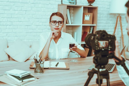 Blogger Makes a Video. Blogger is Upset Businesswoman. Video About a Business. Man Operator Shoots a Video on Camera. Laptop and Supplies on Table. Woman Drinking a Tea. People in Studio Interior.の写真素材