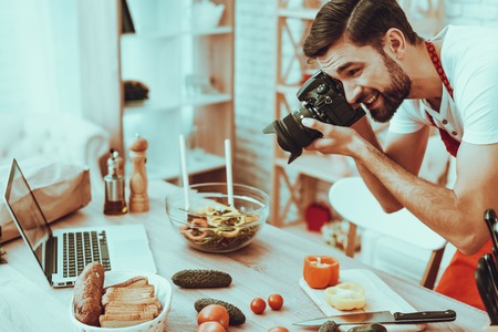 Photographer Makes a Photo. Photographer is Young Beard Man. Man is Taking Picture of Food. Guy is Using a Photo Camera. Laptop and Different Food on Table. Man in Studio Interior.の写真素材