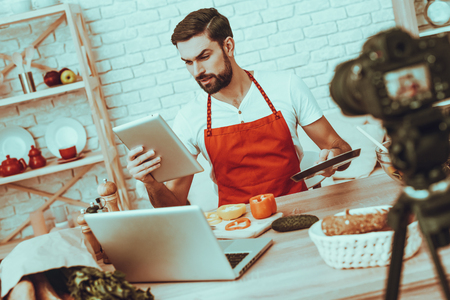 Blogger Makes a Video. Blogger is Smiling Beard Man. Video About a Cooking. Camera Shoots a Video. Laptop and Different Food on Table. Man is Holding a Pan and Tablet PC. Man in Studio Interior.の写真素材