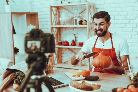 Blogger Makes a Video. Blogger is Smiling Beard Man. Video About a Cooking. Camera Shoots a Video. Laptop and Different Food on Table. Man Showing He Want Cuts a Pumpkin. Man in Studio Interior.の写真素材