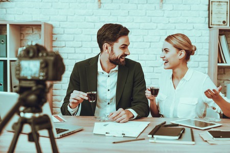 Bloggers Makes a Video. Bloggers is Young Smiling Man and Woman. Video About a Business. Camera Shoots a Video. Gadgets and Supplies on Table. People Drinking a Tea. People in Studio Interior.の写真素材
