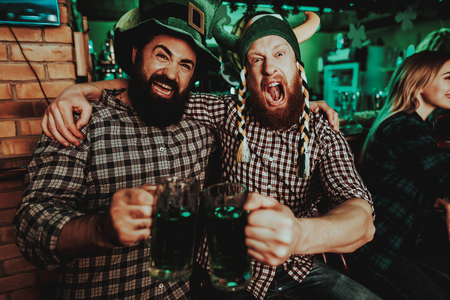 Two Men In Funny Caps. St Patrick's Day Concept. Having Fun. Beer Drinking. Bar Counter. Alcohol Handling. Black Beard. Bearded Males. Good Festive Mood. Bright Lights. Funny Club Visitors.の写真素材