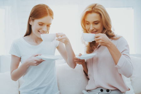 Sitting Mother and Daughter Drinking Tea on Sofa. People in Good Mood. Communication between Parent and Child. Happy Mother and Daughter. Morning Coffe. Smiling Women on White Sofa.の写真素材