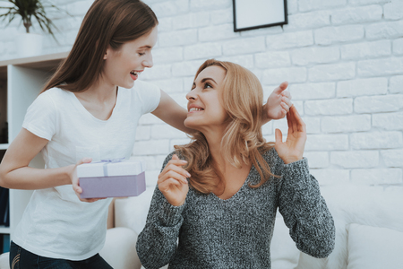 Smiling Daughter Gives Mother Gift in Small Box. Relationship in Family. Holiday at Home. Happy Mother. Friendly Relations. Celebrating Together. Happiness in Family Concept. Presenting Gift.の写真素材