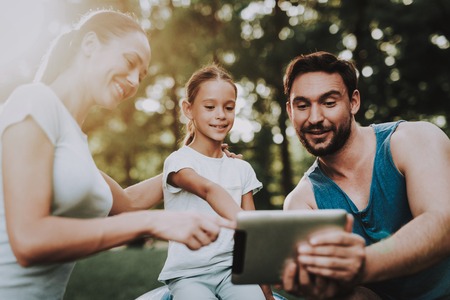Happy Young Family Using Tablet in Summer Park. Sport and Healthcare Concept. Summer Park. Fun Outdoor. Green Grass. Sport in Summer. Father and Mother. Smiling Daughter. Using Digital Device.の写真素材
