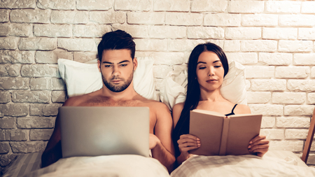 Couple Using Laptop Reading Book Before Sleeping. Young Guy Watching Movie or Browsing Internet. Girl Reading Book at Night. Relationship Concept. Wife and Husband.の写真素材