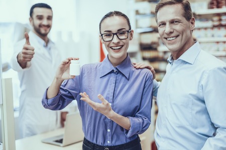 Customers Choose Medicaments. Customers is Adult Man and Young Woman. Woman Showing Bottle with Pills. Pharmacist on Background Showing OK Sign. People is Smiling. People Located in Pharmacy.の写真素材