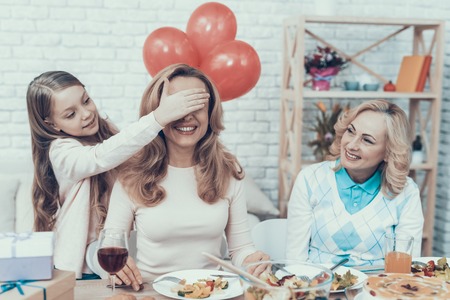 Family Preparing Surprize for Mother at Home. Cake on Table. Happy Family. Mother with Daugther. Smiling Women. Smiling Grandmother. Celebration Concept. Glass of Wine. Closed Eyes. Red Bbaloons.の写真素材