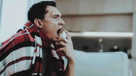 Man with Cold Sitting on Sofa in Checkered Blanket. Man at Home. Sick Young Man. White Sofa in Room. Disease Concept. Healthcare and Healthy Lifestyle Concept. Using Spray. Flu Treatment.の写真素材
