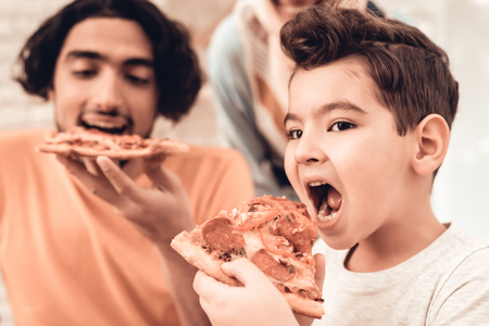 Happy Arabian Family Eating Pizza in Kitchen. Muslim Family. Smiling Boy. Young Arabian Woman. Modern Kitchen at Home. Man Using Kitchenware. Young Family. Wooden Table in Kitchen. Food on Table.の写真素材