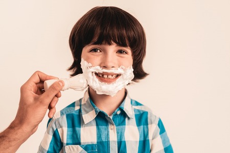 Young Father Teaches Little Son to Shave at Home. Foam on Face. Shaving Tool. Razor with Blade. Happy Family Concept. Young Boy in Shirt. Hygiene and Skincare Concepts. Hygienic Soap.の写真素材