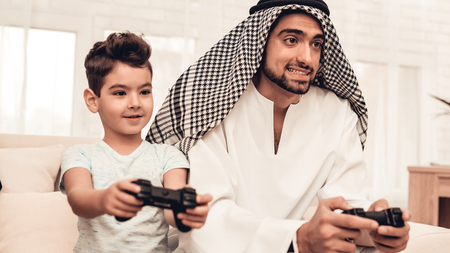 Happy Arabian Family Playing on Console at Home. Family Sitting on Sofa. Man Using Digital Device. Smiling Boay at Home. Muslim Family. Smiling Boy. Young Arabian Woman. Woman in Black Dress.の写真素材