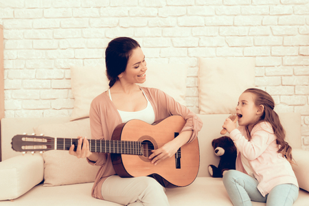 Kids Home Games. Rest at Home. Child Development. Mom and Daughter Play. Happy Mom and Child in Pajamas. Woman and Daughter. Play Guitar. Woman Playing Guitar. Mother Teaches Daughter. White Interiorの写真素材