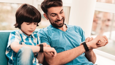 Little Boy with Father Wacthing on Hand Watches. Sitting Boy. Young Father. Young Bearded Man at Home. Sitting on Blue Sofa. Happy Family Concept. Time Concepts. Modern Hand Watch.の写真素材