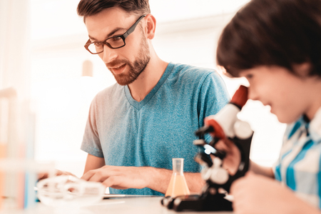 Youn Bearded Father Teaching Son in Shirt at Home. Education at Home. Using Microscope. Studding Chemistry. White Table in Room. Sitting Boy. Young Father. Lesson at Home. Education Concept.の写真素材