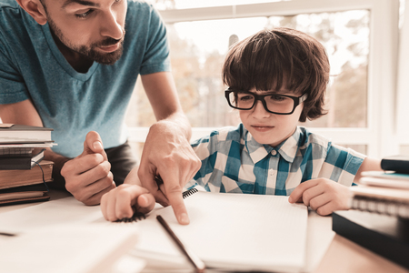 Little Boy in Glasses Doing Homework with Father. Education at Home. White Table in Room. Sitting Boy. Young Father. Lesson at Home. Education Concept. Writing in Notebook. Boy in Shirt.の写真素材