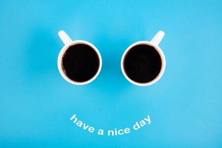 Two white coffee mugs with a smile on a blue background with the phrase Have s nice day. Happy coffee mugs.の写真素材