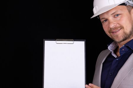 Engineer, builder, businessman in a white helmet on a dark background holds a white tablet in his...