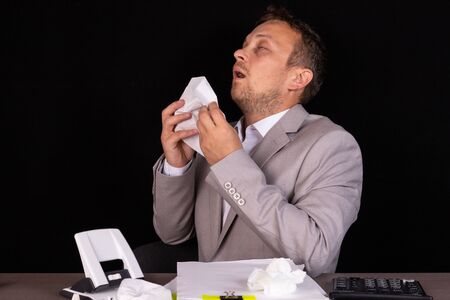 Portrait of a sick entrepreneur blowing in a napkin in an office with used napkins on a tableの写真素材