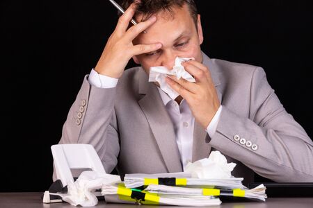 Portrait of a sick entrepreneur blowing in a napkin in an office with used napkins on a tableの写真素材