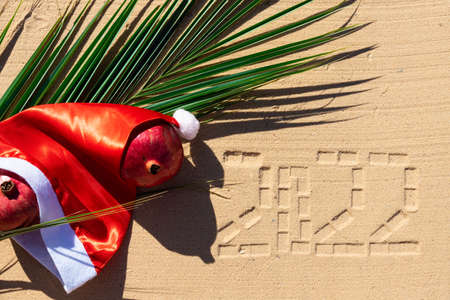 Hat of Santa Claus on the background of a palm leaf, with pomegranate fruits on the sandy beach of the Red Sea. Tropical Christmas card.の写真素材