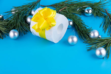 A roll of toilet paper as a Christmas present near a branch of a Christmas tree on a blue...