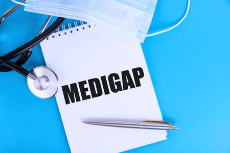 MEDIGAP, text written in a notebook lying on a blue background, with a stethoscope and a medical mask. Medical concept.の写真素材