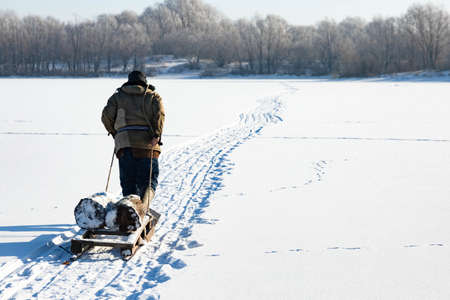 A lonely old hermit, in winter, crosses the river on ice and pulls a sled with firewood for heatingの写真素材