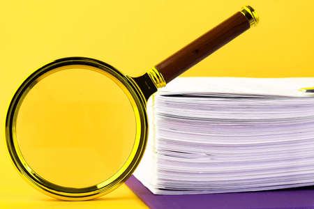 Close up of a pile of unfinished documents awaiting verification, a magnifying glass, on a yellow background. Business and education concept. Copy spaceの写真素材