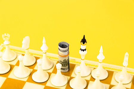 Close-up of rolled US one hundred dollar bills standing on a chessboard instead of a chess piece. Yellow background. Copy space.の写真素材