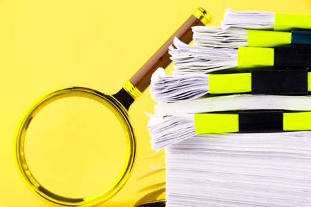 A large stack of unfinished documents awaiting verification, a magnifying glass, against a yellow background. Business and education concept. Copy spaceの写真素材