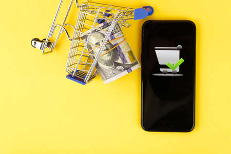Online shopping, telephone and shopper trolley concept with roll of one hundred dollar bills on yellow background. Copy space.の写真素材