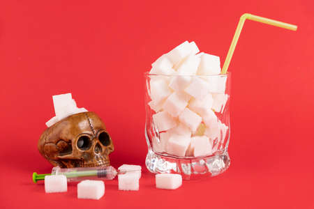 A glass of white glass with a straw, filled with white cubes of refined sugar and a human skull on a red background. Copy space.の写真素材
